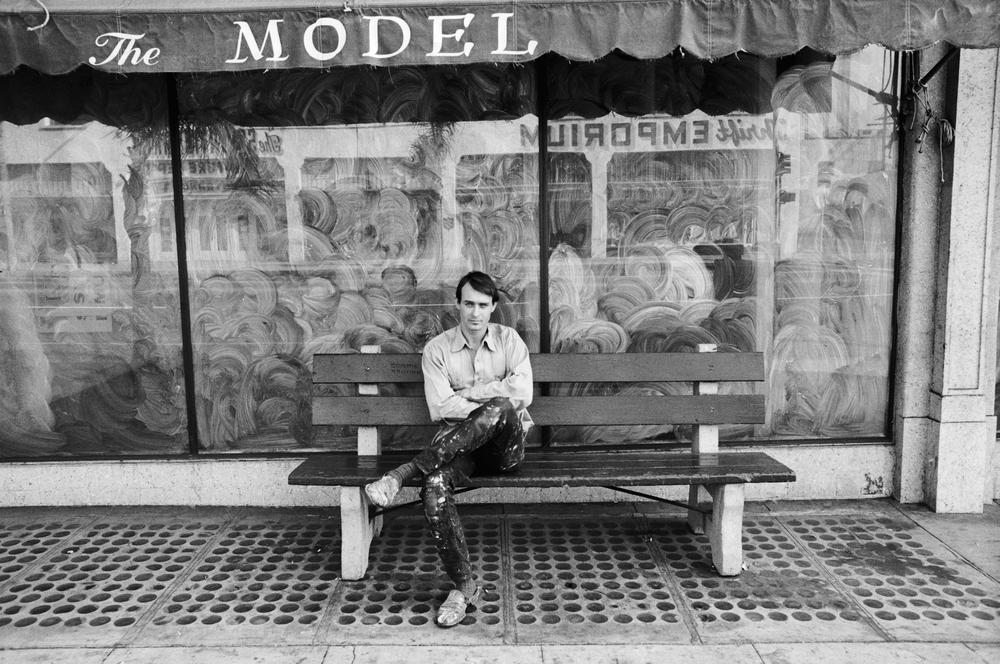 artist sitting with one leg crossed on a bench outside of a storefront's glass window