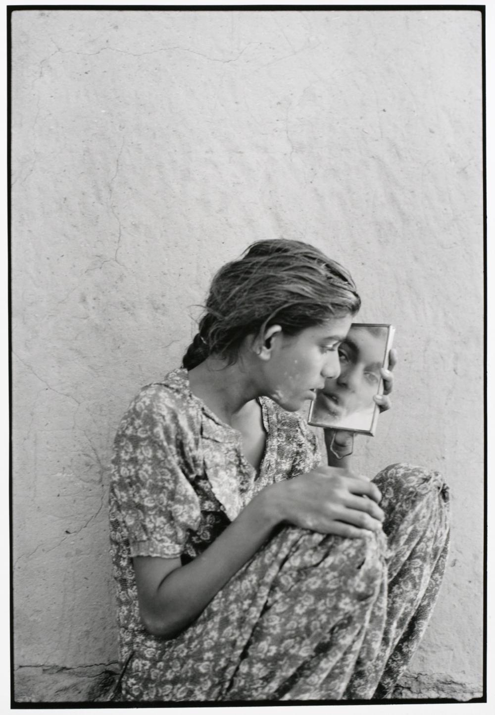 woman in patterned dress sitting against a white wall and looking very closely in an angled mirror so that the viewer can see both her profile and reflection